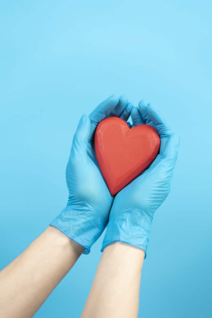Female hands wearing blue medical gloves holding red heart over light blue background. Health care, heart disease, cardiology